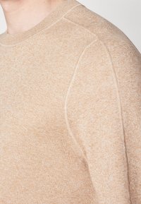 Gros plan sur une personne portant un pull en tricot beige clair, à texture douce, avec des détails de coutures visibles sur l'épaule et la partie supérieure du bras.
