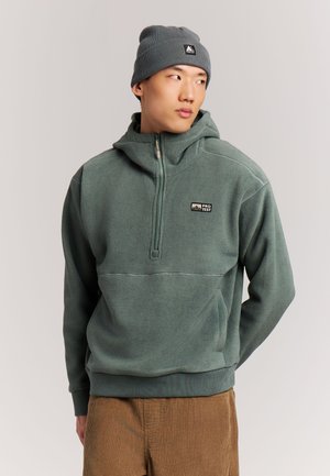 Junger Mann trägt einen grünen Fleecepullover mit halbem Reißverschluss und eine graue Mütze, schaut nach links mit den Händen hinter dem Rücken.