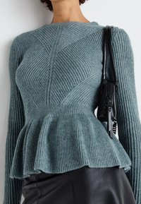 Femme portant un pull en tricot côtelé teal avec un ourlet peplum, une jupe en cuir noire et un sac à bandoulière noir sur fond uni.