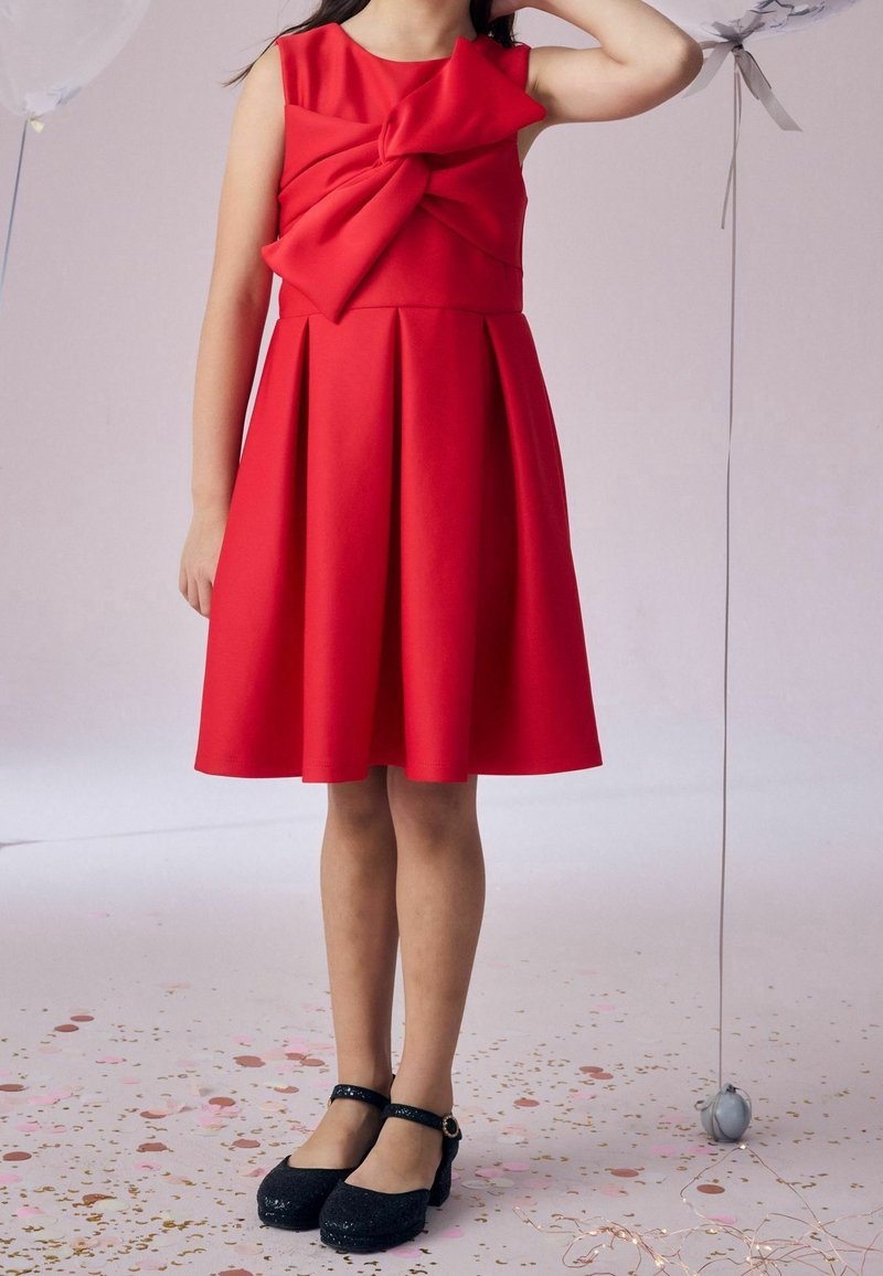 Enfant portant une robe rouge sans manches avec un grand nœud sur la poitrine et des chaussures noires à brides à la cheville, debout sur un sol couvert de confettis à côté d’un ballon.
