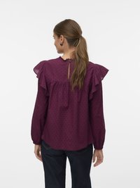 Femme avec une queue de cheval brune portant un blouse violette à manches longues avec des volants et de petits points texturés, assortie à un jean foncé, de dos.