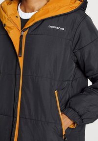 Chaqueta acolchada negra con capucha y detalles en naranja contrastante. Incluye cremalleras, bolsillos laterales y un tejido texturizado. Logo de la marca visible.