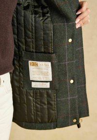 Interno della giacca tweed con fodera trapuntata di colore verde scuro. Presenta un'etichetta con il marchio e le istruzioni per la cura. Sono presenti caratteristici bottoni di chiusura.