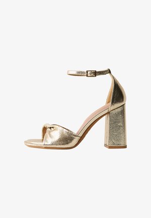 Gold metallic open-toe sandal with block heel and ankle strap with buckle, shown from the side on a white background.