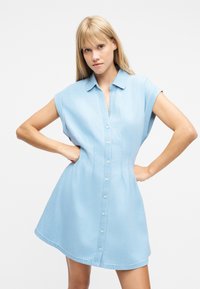 Γυναίκα με ίσια ξανθά μαλλιά, φορούσε ανοιχτό μπλε φόρεμα από τζιν χωρίς μανίκια με κουμπιά, στέκεται με τα χέρια στη μέση μπροστά σε λευκό φόντο.