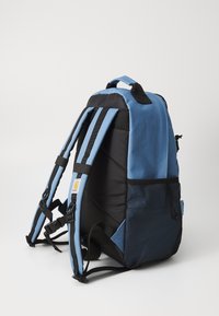 Blauer und schwarzer Stoff-Rucksack mit gepolsterten Trägern, Netzseitentasche, Tragegriff oben und Logo-Patch auf der Vorderseite. Seitenansicht.