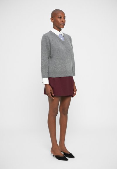 Grauer Strickpullover mit V-Ausschnitt, getragen über einem weißen Hemd mit blauen Streifen, kombiniert mit einem burgunderfarbenen Rock und schwarzen Absatzmules.