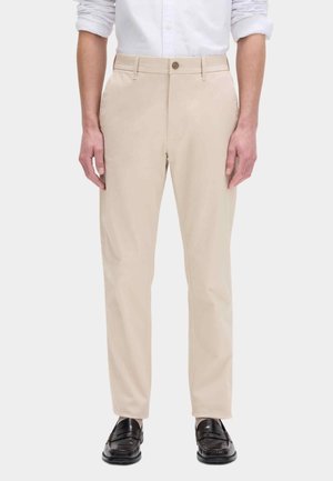 Mand iført beige bukser med lige ben, hvid skjorte med lange ærmer med opkørte ærmer og sorte læder loafers, der står mod en ensfarvet baggrund.