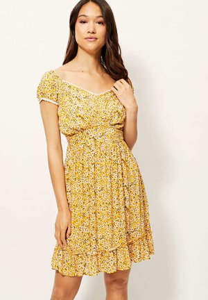 Mujer con cabello largo y castaño que lleva un vestido corto amarillo con estampado floral, mangas abullonadas y bajo con volantes, de pie frente a un fondo liso.