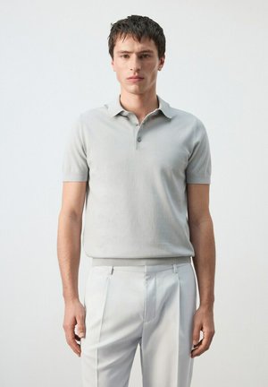 Jeune homme debout face à l'avant portant un polo à manches courtes gris clair et un pantalon plissé blanc sur un fond uni.