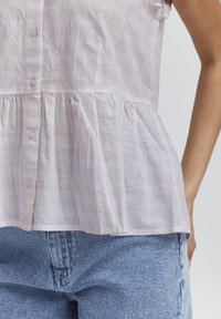 Blouse rose clair texturée avec un ourlet froncé, dotée d'un devant à boutons et de manches courtes. Assortie à un jean en denim bleu taille haute.