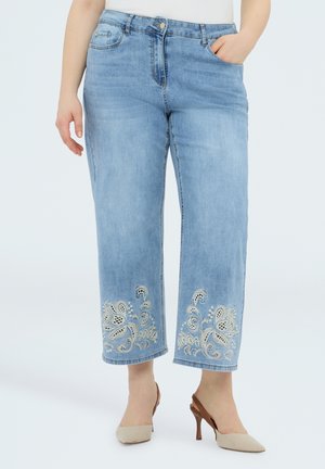 Hellblaue kurze Jeans mit Blumenstickerei am Saum, kombiniert mit beigefarbenen Slingback-Pumps, Modell steht mit der Hand in der Tasche.
