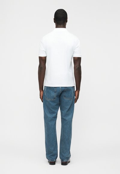 Homme debout, dos tourné, portant un polo blanc à manches courtes et un jean bleu ample, devant un fond clair uni.