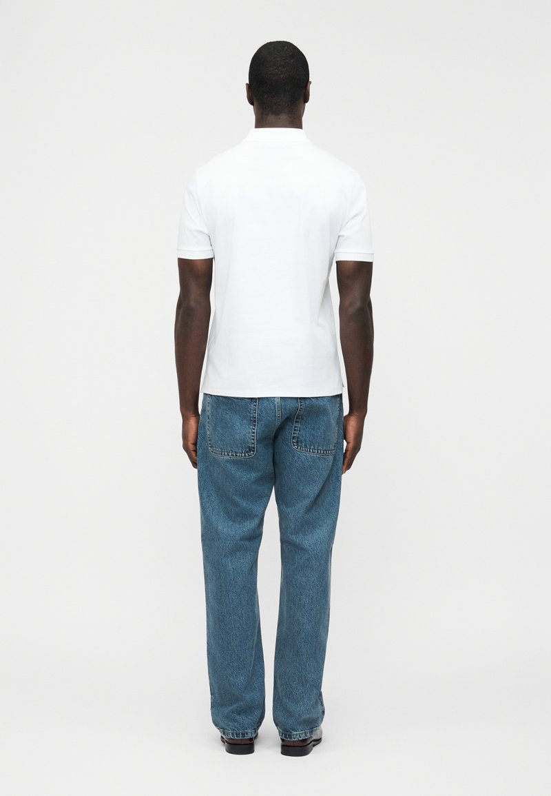 Man standing facing away, wearing a white short-sleeve polo shirt and loose blue jeans against a plain light background.