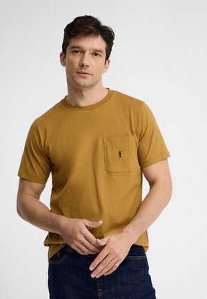 Mann trägt ein senfgelbes kurzärmeliges T-Shirt mit kleinem Golfspieler-Logo auf der Brusttasche, steht vor einfachem hellem Hintergrund.