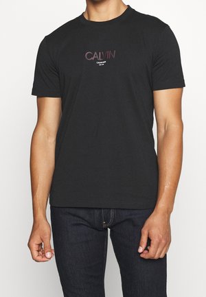 Homme portant un T-shirt Calvin Klein noir à manches courtes et un jean foncé, debout devant un fond blanc uni.