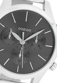 Montre en métal argenté avec cadran gris, trois sous-cadrans, index des heures argentés et bracelet en maillage. Présente une finition lisse et un design clair et lisible.