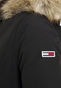 Veste noire à finition texturée, avec un col en fausse fourrure et une étiquette blanche ornée de rayures horizontales rouge, blanc et bleu.