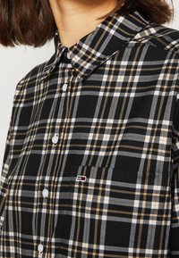 Camicia a quadri in bianco e nero con accenti marroni e grigi, dotata di colletto e bottoni. Include una piccola etichetta con logo sul petto.
