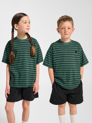 To børn iført matchende grønne og sorte stribede t-shirts og sorte shorts står side om side mod en ensfarvet baggrund.