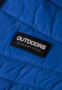 Mėlynos pūkinės striukės audinys su siūlių raštu. Yra juodas stačiakampis etiketė su baltu tekstu: "OUTDOORS Expedition."