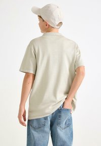 Ljusgrå kortärmad polo med en avslappnad passform, kombinerad med blå denimjeans. Bakgrunden är enfärgad vit för att lyfta fram outfiten.