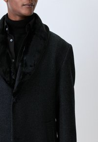 Čierny károvaný kabát s mäkkým čiernym kožušinovým golierom, s klopovým golierom, dvoma gombíkmi a textúrovanou tkaninou. Skryté vrecká po stranách.