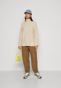 Pulover bej cu guler înalt, pantaloni cargo maro cu cusături, șapcă din denim, geantă galbenă și adidași argintii. Design simplu, relaxat.