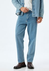 Jeans in denim azzurro chiaro con taglio dritto, design classico a cinque tasche e zip frontale. Indossati con una giacca in denim azzurro chiaro e stivali marroni.