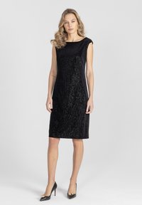 Czarna sukienka z cekinami o dopasowanym kroju, bez rękawów, z okrągłym dekoltem, zestawiona z czarnymi, szpiczastymi szpilkami ozdobionymi złotymi detalami.