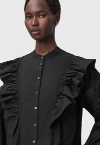 Blusa nera con maniche a volant, chiusura con bottoni e cuciture verticali. Realizzata in tessuto leggero con una finitura liscia.