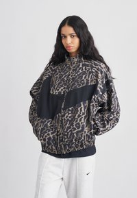 Μπουφάν bomber με μοτίβο λεοπάρ σε μπεζ και μαύρο, με ψηλό γιακά, κλείσιμο με φερμουάρ και ελαστική ρίγα στο κάτω μέρος. Συνδυασμένο με ανοιχτό γκρι φόρμες.