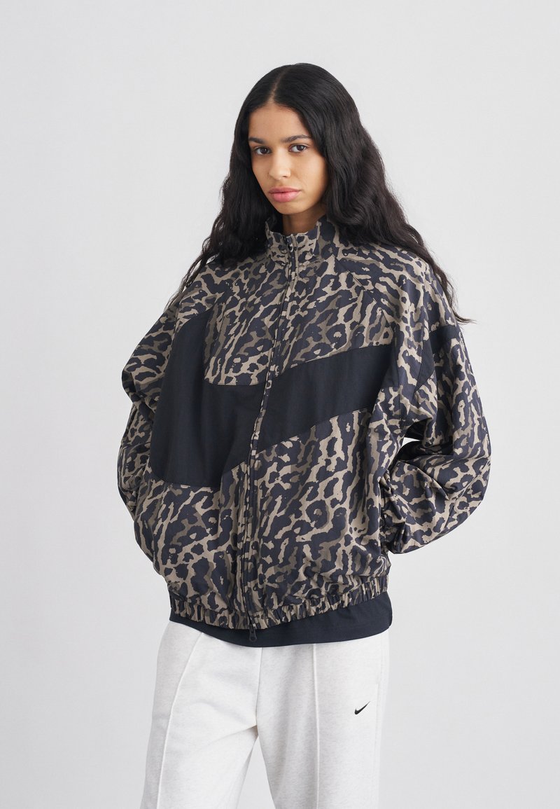 Μπουφάν bomber με μοτίβο λεοπάρ σε μπεζ και μαύρο, με ψηλό γιακά, κλείσιμο με φερμουάρ και ελαστική ρίγα στο κάτω μέρος. Συνδυασμένο με ανοιχτό γκρι φόρμες.