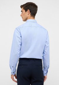 Camicia azzurro chiaro a maniche lunghe con colletto e polsini abbottonati, abbinata a pantaloni blu navy con due tasche posteriori e passanti per cintura.