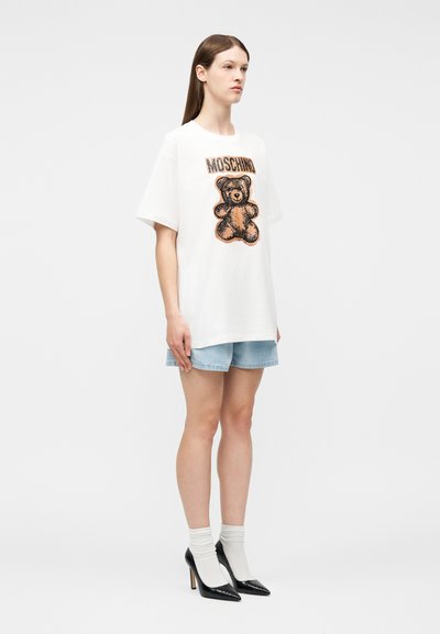 Γυναίκα που στέκεται φορώντας υπερμεγέθη λευκό T-shirt με αρκουδάκι και το κείμενο "MOSCHINO", ανοιχτόχρωμα τζιν σορτς, λευκές κάλτσες και μαύρες γόβες.