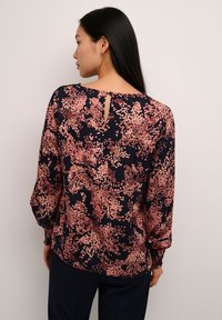 Blouse à manches longues en tissu bleu marine avec un motif floral aux nuances de rose et de pêche. Présente un détail œillet au dos du col.
