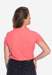 Koraalkleurig polo shirt met een kraag, korte mouwen en een achterste plooi. Draagt men met hoge taille zwarte jeans, die een gladde textuur tonen.