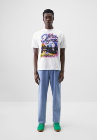 Hvit bomull t-skjorte med fargerik "Amsterdam" grafikk, sammen med lyseblå mønstrede bukser og grønne sneakers med brune såler.