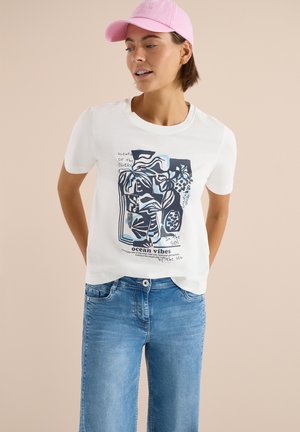 Frau trägt ein weißes T-Shirt mit blauem, ozeanischem Grafikdesign, hellblaue Jeans und eine pinke Baseballkappe, steht vor einem beigen Hintergrund.