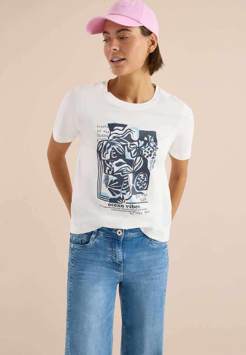 Femme portant un t-shirt blanc avec un motif graphique bleu sur le thème de l'océan, un jean bleu clair et une casquette de baseball rose, debout devant un fond beige.