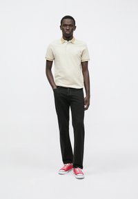 Lys beige polo-shirt med gul krage, kombinert med svarte jeans og røde sneakers. Modellen står med den ene hånden i lomma foran en hvit bakgrunn.