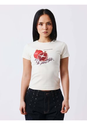 Jeune femme aux cheveux raides et foncés portant un t-shirt blanc moulant avec un motif de grenade rouge et un pantalon taille haute foncé.