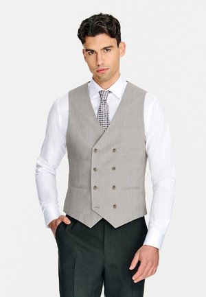 Mężczyzna w jasnoszarej dwurzędowej kamizelce, białej koszuli, wzorzystym krawacie i ciemnych spodniach stojący na jednolitym tle.