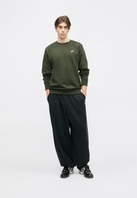 Sweatshirt verde escuro com decote redondo e pequeno logótipo rosa, combinado com calças largas pretas e sapatilhas de desporto pretas.