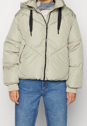 Personne portant une doudoune matelassée beige clair avec des cordons noirs, fermeture éclair devant et jean bleu sur un fond uni.