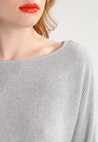 Ljust grå stickad tröja med vid halsringning och ribbat mönster. Har avslappnad passform och mjukt, slätt tyg.