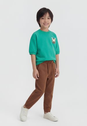 Παιδί που στέκεται και χαμογελά, φοράει πράσινο πουκάμισο με κηλίδα ζέβρας και καφέ παντελόνι, ποζάροντας σε λευκό φόντο.