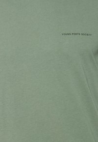 Zelené bavlněné tričko s kulatým výstřihem a krátkými rukávy, s nápisem "YOUNG POETS SOCIETY" černě vytištěným na přední straně.