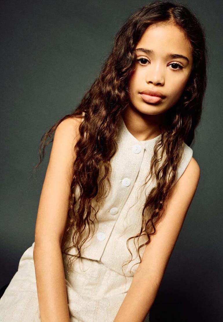 Jeune fille aux longs cheveux bruns bouclés portant un haut blanc sans manches à boutons, sur un fond gris foncé.