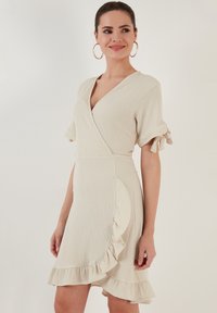Cream-colored wrap dress with short sleeves, featuring ruffle accents along the hem and sleeves, made of textured lightweight fabric.